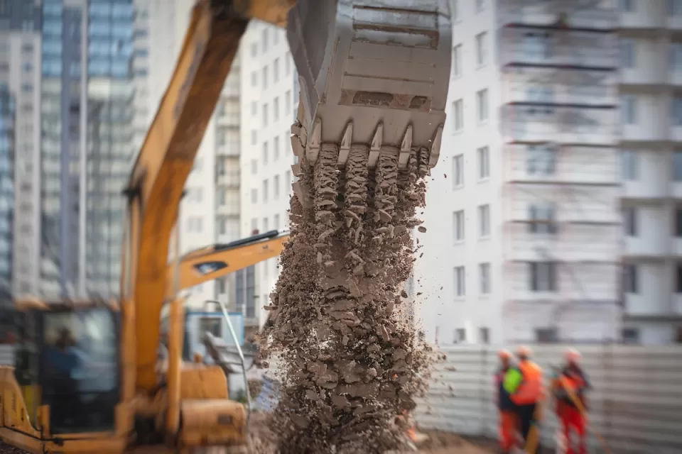 Excavator digging in an urban street with two workers standing next to it.